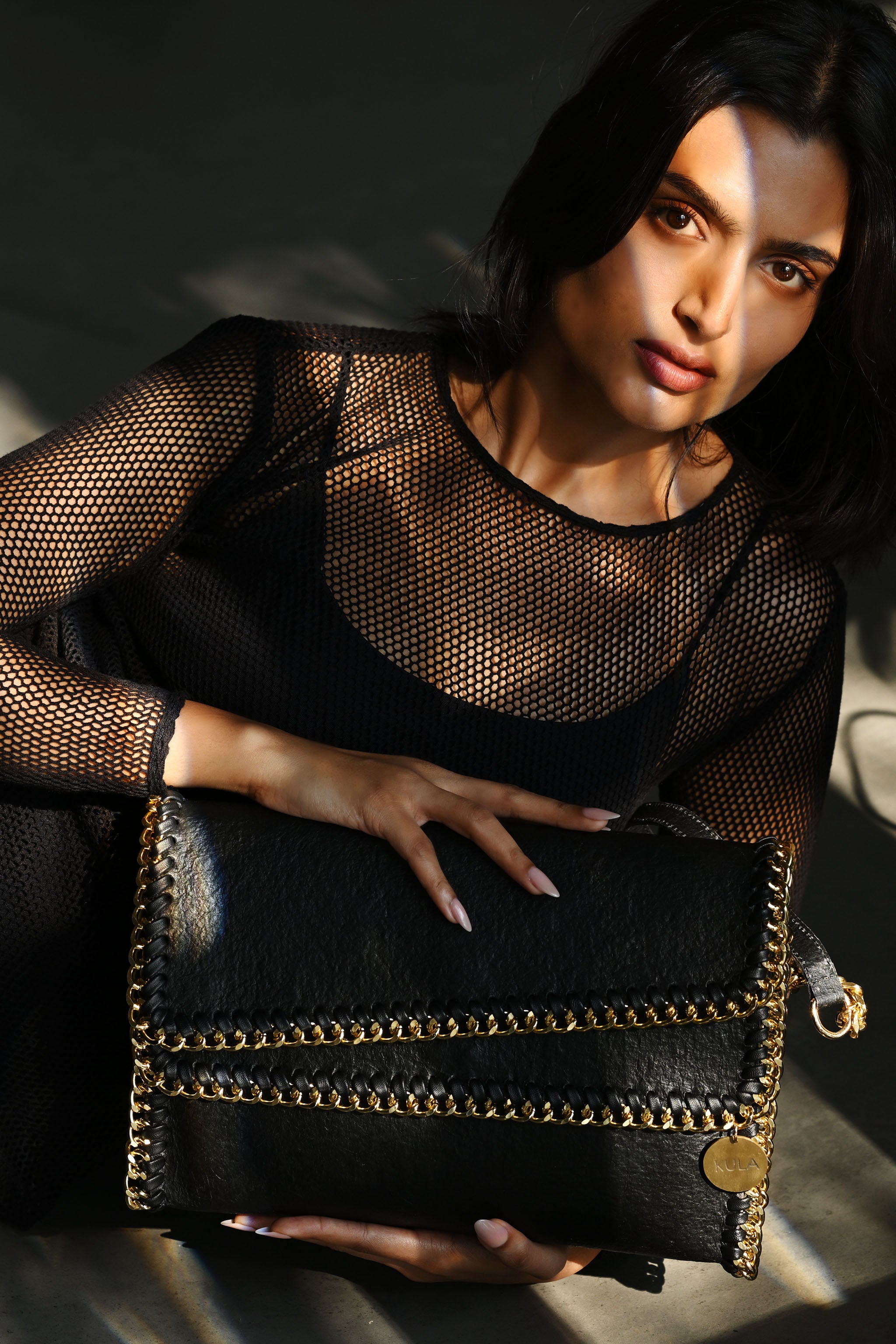 Female model holding a charcoal black bella handbag with gold accents against a dark background